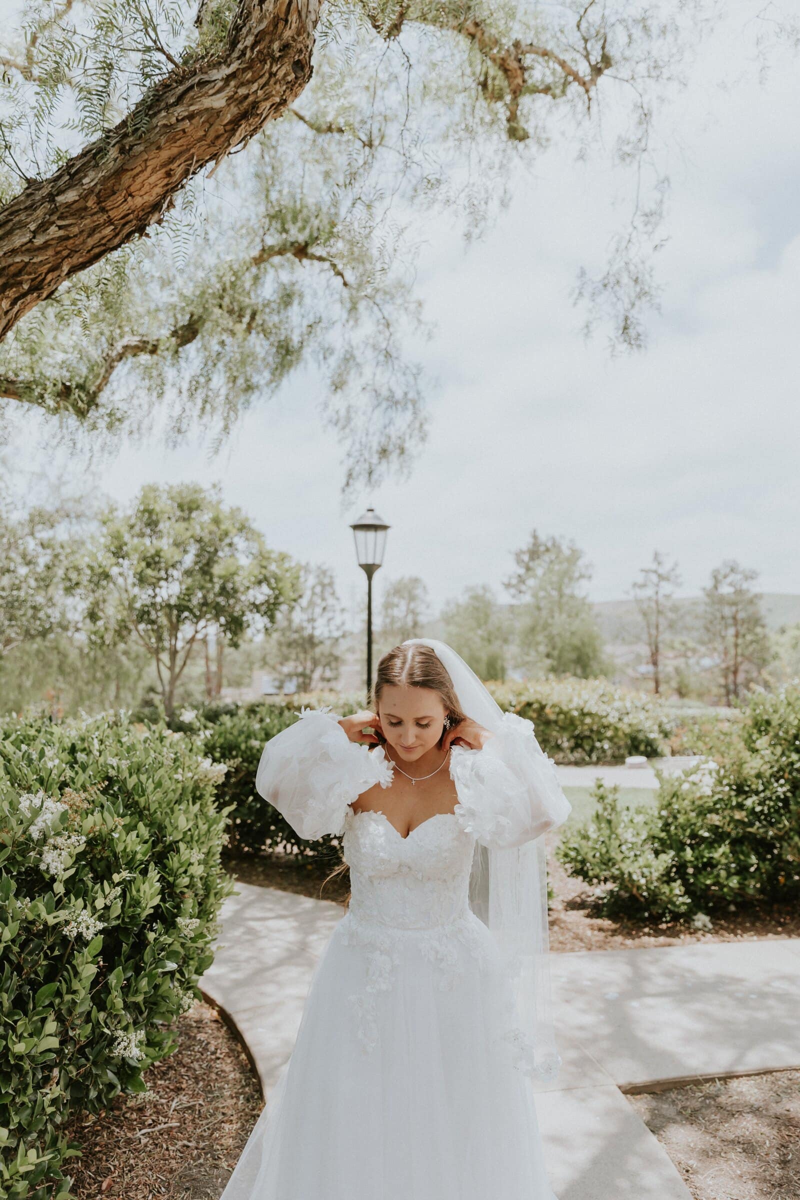 Irelyn on a garden path under a pepper tree, adjusting her earring