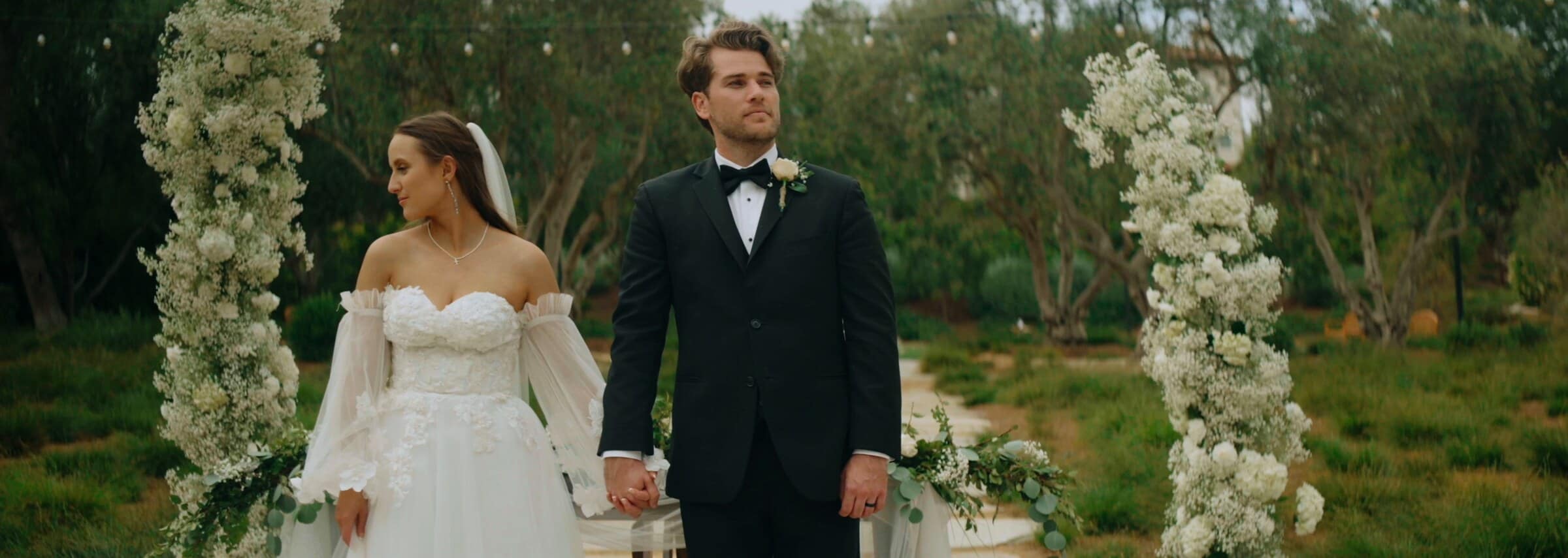 Irelyn and Aleck holding hands between two columns of white flowers