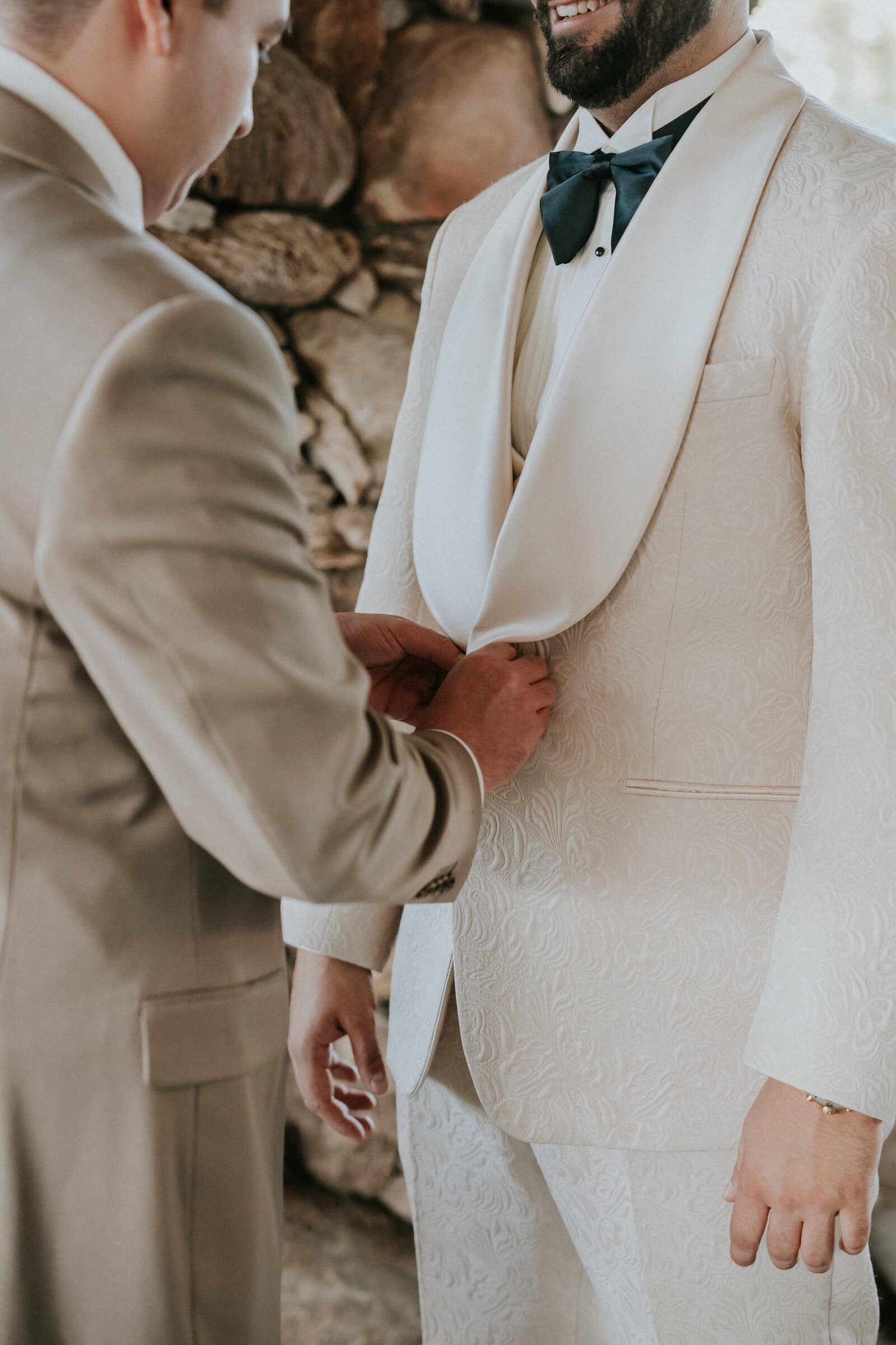 A groomsman buttoning Dom's cream tuxedo jacket