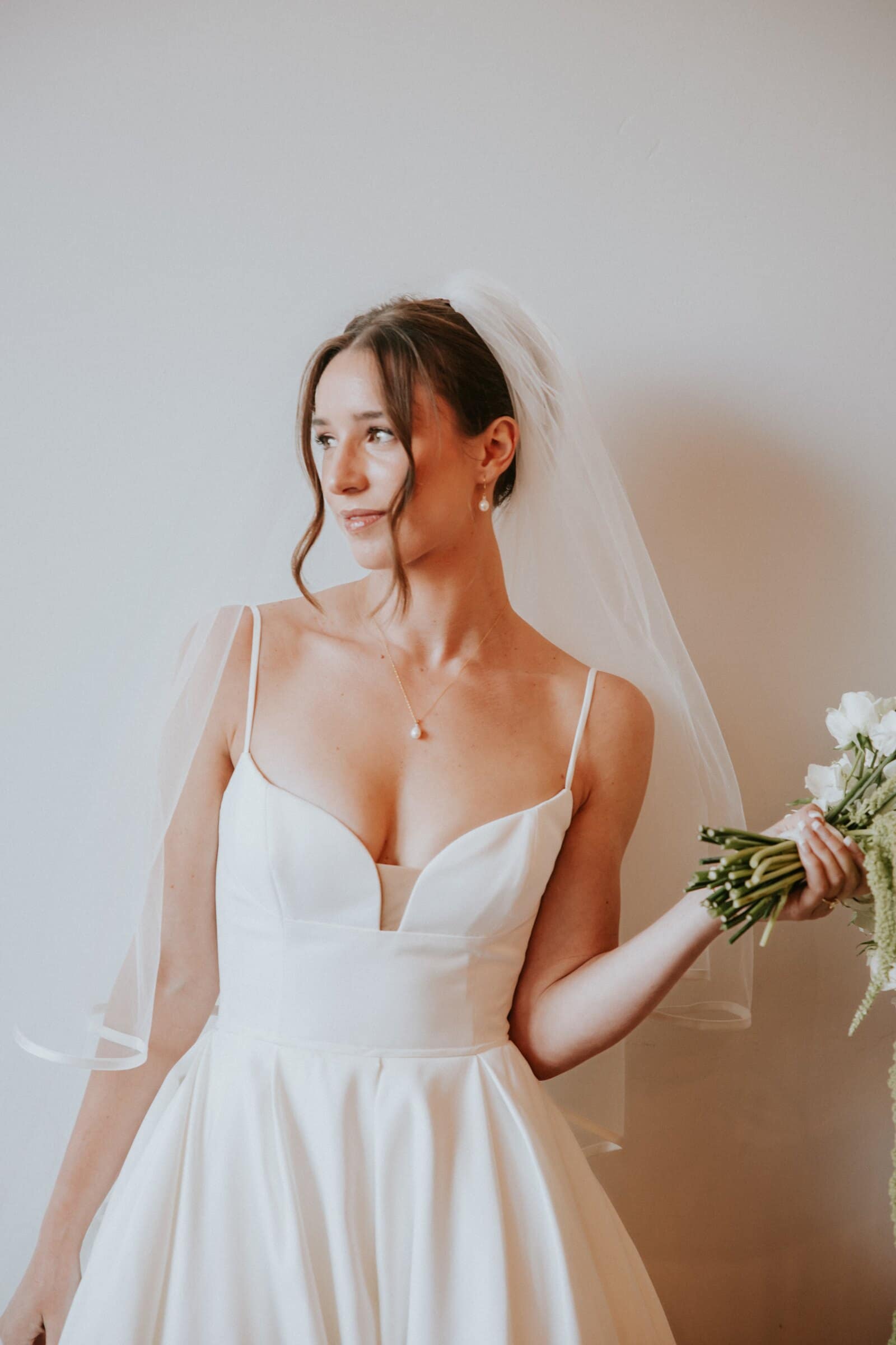 Clara in her veil against a white wall, bouquet in hand