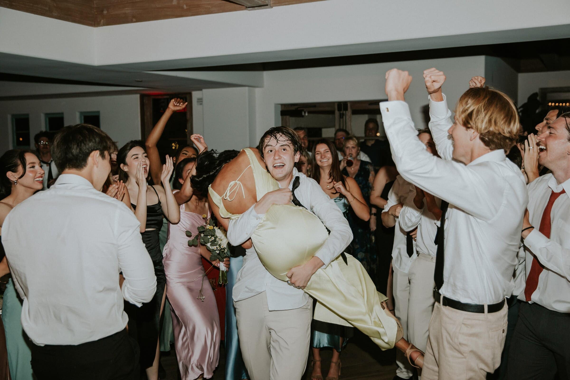 A groomsman carrying a bridesmaid across the dance floor as friends cheer